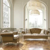 Luxury Italian furniture in elegant living room setting with brown tufted sofa and armchair, large windows, and chandeliers.