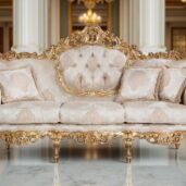 A luxurious sofa with golden details and floral beige upholstery, a tufted backrest and decorative cushions, set in an elegant space with marble flooring and columns.