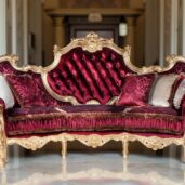 A luxurious sofa featuring red velvet upholstery and golden accents, a high tufted backrest, and decorative cushions, set within an elegant room adorned with classical columns and stucco work.