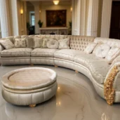 Divano curvo di lusso con dettagli dorati, tessuto chiaro decorato e cuscini eleganti in un atrio con colonne e pavimento in marmo.