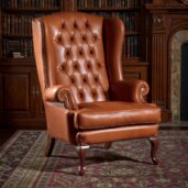 Poltrona wingback in pelle color cognac, schienale capitonné, braccioli arrotolati, profilo con borchie e gambe a cabriole in legno.