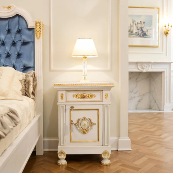 Comodino di lusso laccato avorio con dettagli in foglia oro, top profilato, cassetto e anta con medaglione scolpito, maniglieria in ottone e piedi lavorati, accanto a letto con testiera in velluto blu.