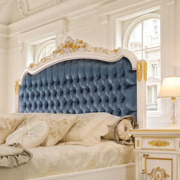 Letto di lusso laccato avorio con dettagli dorati, testiera capitonné in velluto blu, panchetta coordinata e comodini neoclassici in elegante camera.