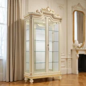 Luxury ivory-lacquered display cabinet with gold accents, glass front, glass shelves and a carved crown on turned feet in an elegant classic interior.