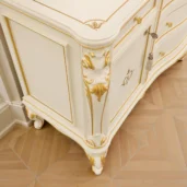 Close-up of a classic ivory-lacquered dresser with gold accents: hand-carved acanthus corner, sculpted foot and curved drawer front with brass knobs.
