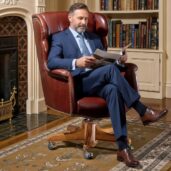 Uomo professionale seduto su una poltrona direzionale girevole in pelle marrone di lusso, leggendo un libro in relax davanti a un camino, in un salotto executive con librerie, ideale per arredi di alta gamma e comfort ergonomico.