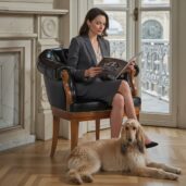 Donna elegante seduta su una poltroncina direzionale in pelle nera di lusso, leggendo un libro in un salotto luminoso con camino e finestre, ideale per arredi home office di alta gamma e comfort sofisticato.