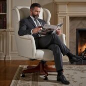 Uomo elegante seduto su una poltrona direzionale girevole in pelle crema di lusso, leggendo una rivista con un bicchiere in mano davanti a un camino acceso, in un salotto executive con librerie, ideale per arredi di alta gamma e relax professionale.