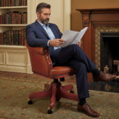 Uomo seduto su poltrona presidenziale EISENHOWER in pelle marrone, in uno studio classico con camino e libreria — Marzorati