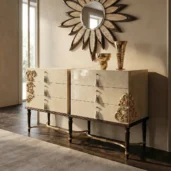 Роскошный комод на 6 ящиков с глянцевой кремовой лакировкой, украшенный золотой цветочной резьбой по углам, тканевыми ручками-кистями и ножками из темного дерева с золотыми наконечниками.