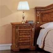Elegant handcrafted Regina wooden nightstand with ornate details next to bed, topped with silver lamp and books.
